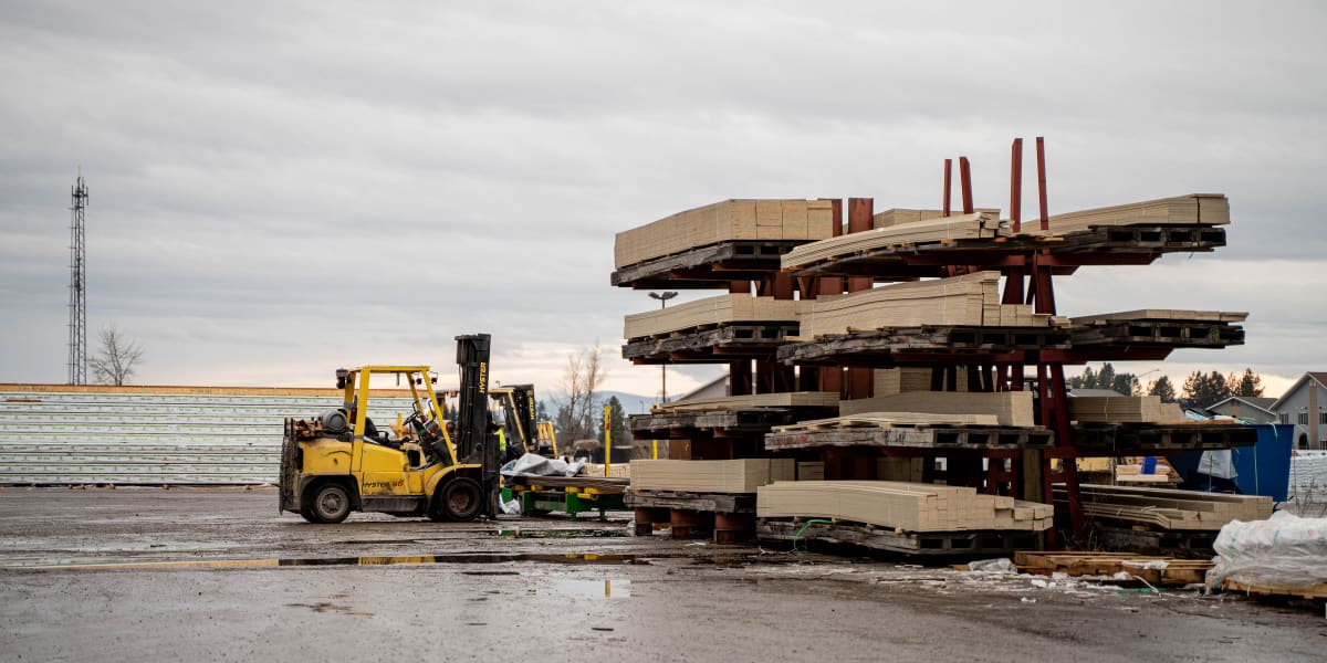A lumber yard storage area showing stacked engineered lumber beams and building materials organized on red metal storage rack