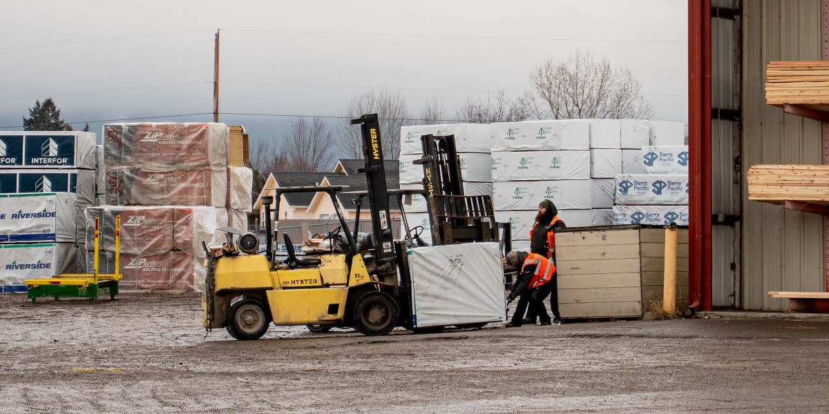 A lumber yard scene showing a yellow Hyster forklift moving wrapped building materials with a worker in an orange safety vest