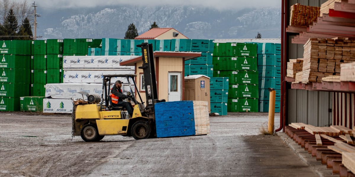 A lumber yard warehouse facility with a yellow Hyster forklift carrying a blue pallet in the foreground