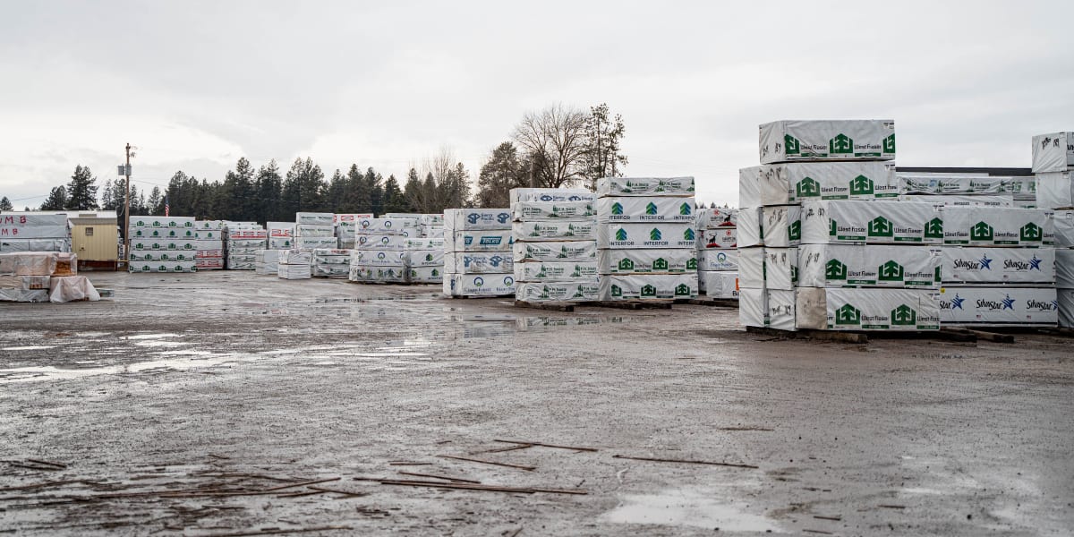A lumber yard storage area with numerous pallets of wrapped building materials stacked in organized rows across a wet concret