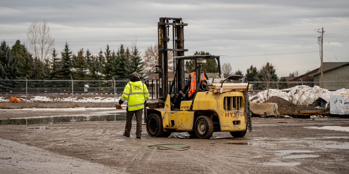 A construction jobsite scene showing a yellow Hyster forklift being operated by workers in high-visibility safety vests