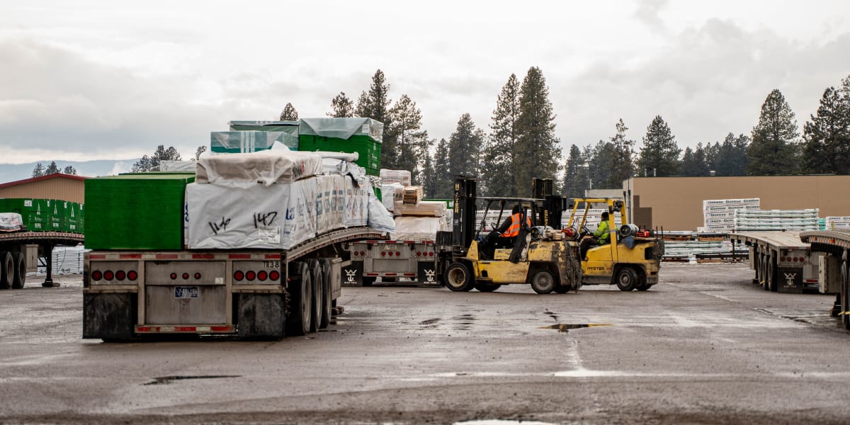 Industrial lumber yard scene showing flatbed trucks being loaded with building materials by yellow forklifts
