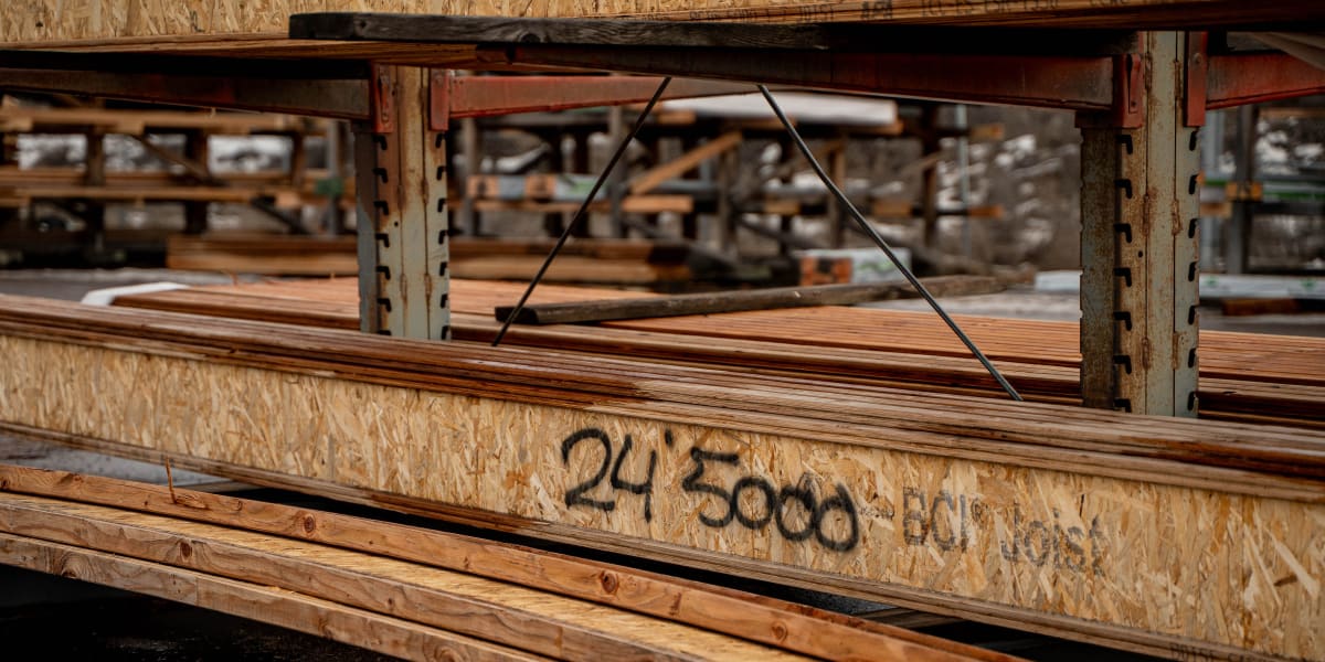 A lumber yard storage area showing wooden engineered lumber beams or I-joists stacked on metal racks