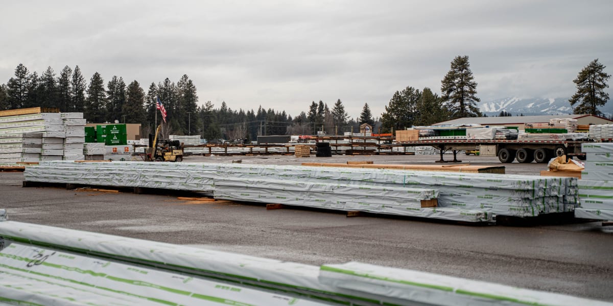 A lumber yard facility showing organized stacks of building materials including wrapped lumber bundles, dimensional lumber, a