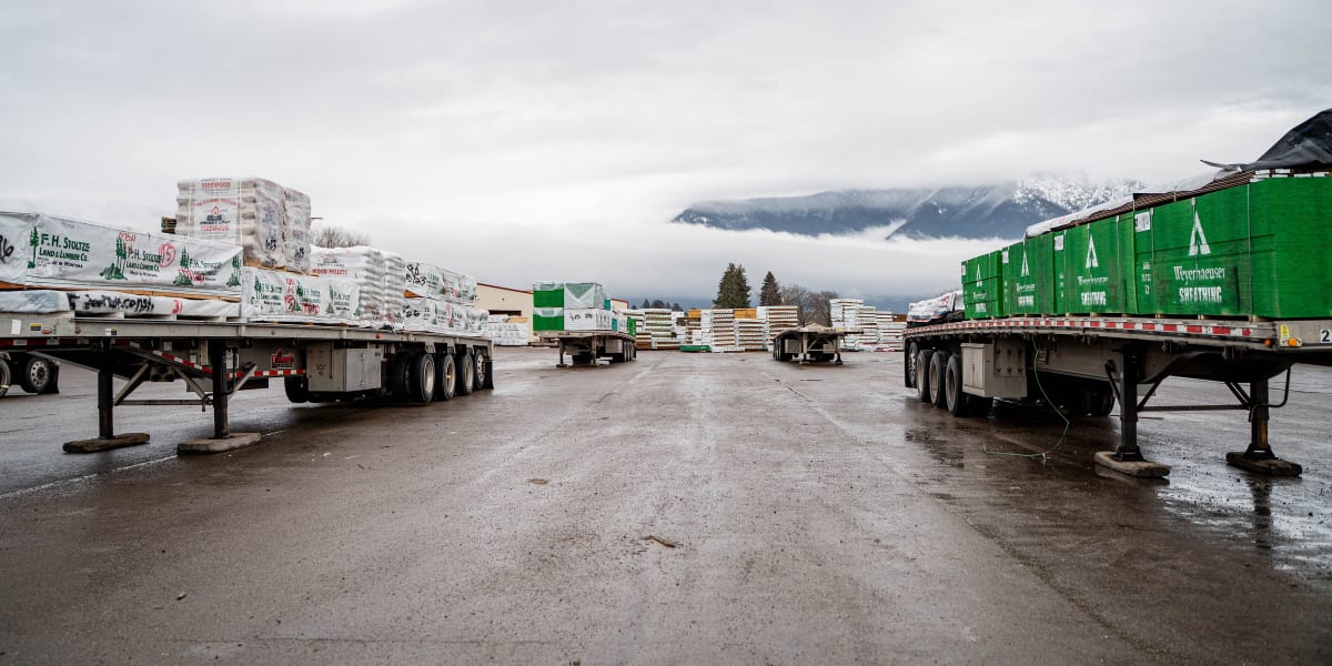 A wide view of a building supply yard showing two flatbed semi-trailers loaded with building materials positioned on either s