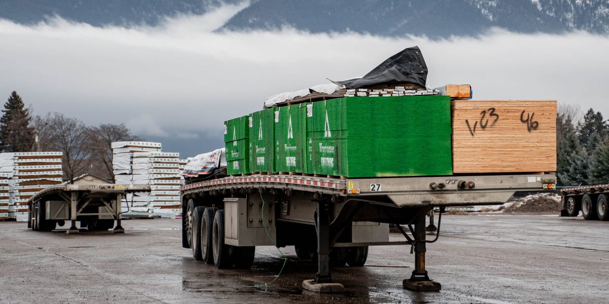 A semi-trailer loaded with green shipping containers and wooden crates is parked at an industrial yard facility