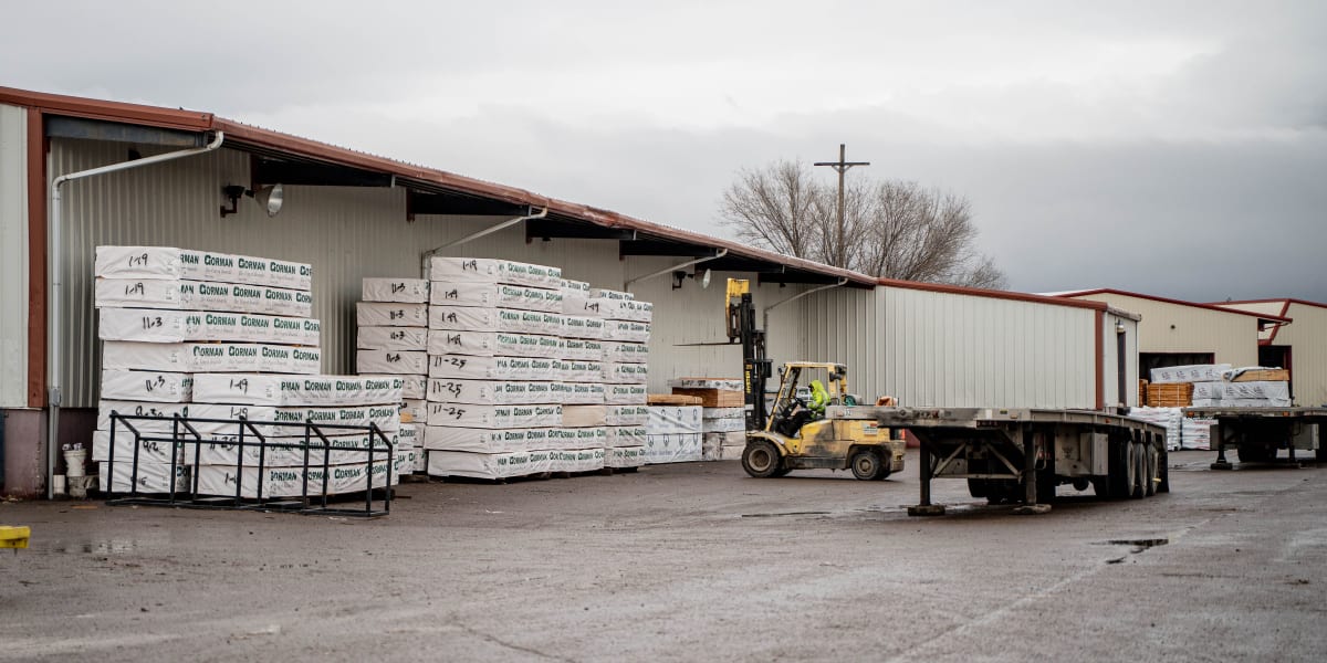 A lumber yard loading dock scene showing stacks of wrapped lumber bundles with handwritten labels stored under an open wareho