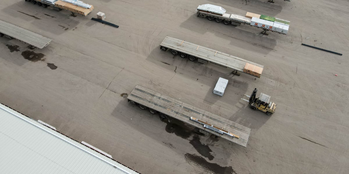 Aerial view of a corporate lumber yard showing multiple flatbed semi-trailers loaded with building materials including metal