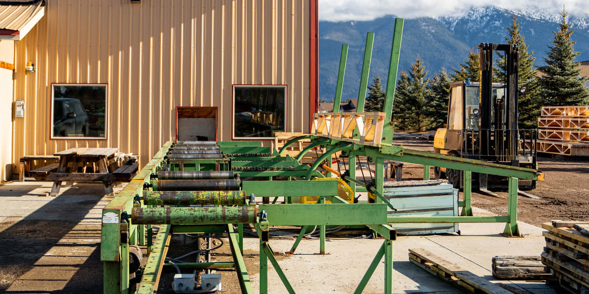 Industrial lumber processing facility showing a large green sawmill or planer machine positioned outside a metal building wit