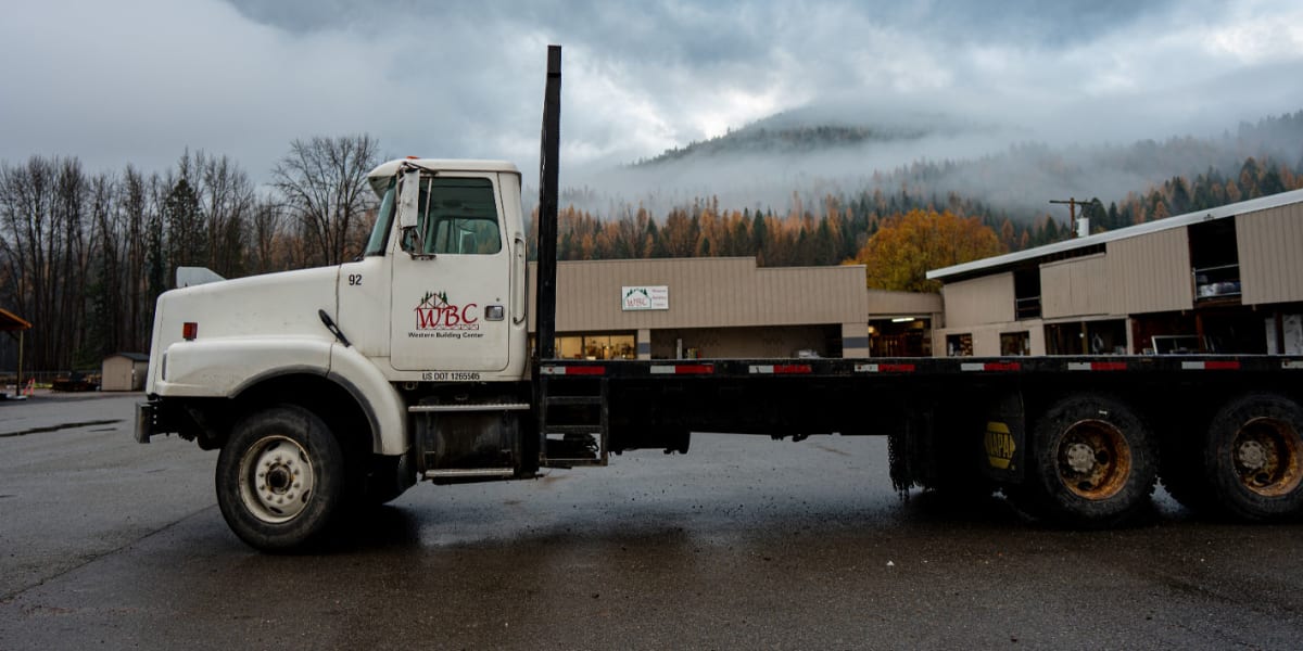 A white flatbed delivery truck with 'WBC' (Western Building Center) branding is parked in front of a large tan-colored buildi