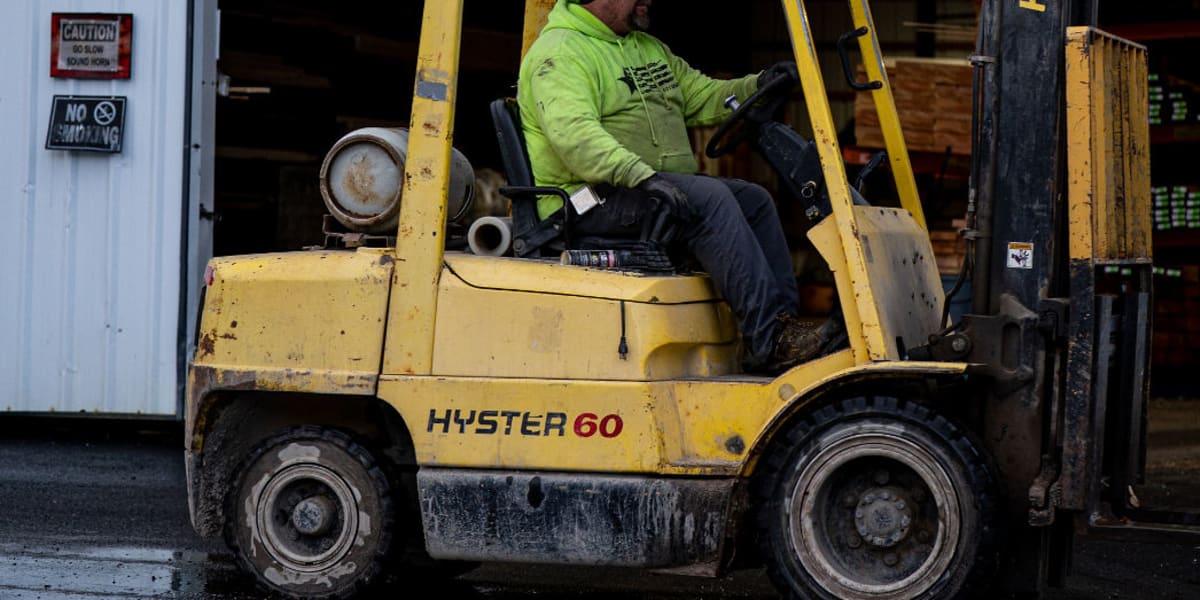 A worker in a bright green safety jacket operates a yellow Hyster 60 forklift outside a metal warehouse building