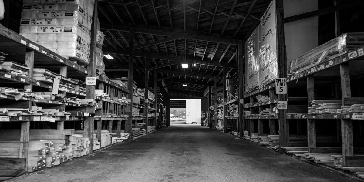 Black and white photograph of a lumber warehouse interior showing tall stacks of dimensional lumber organized on wooden shelv