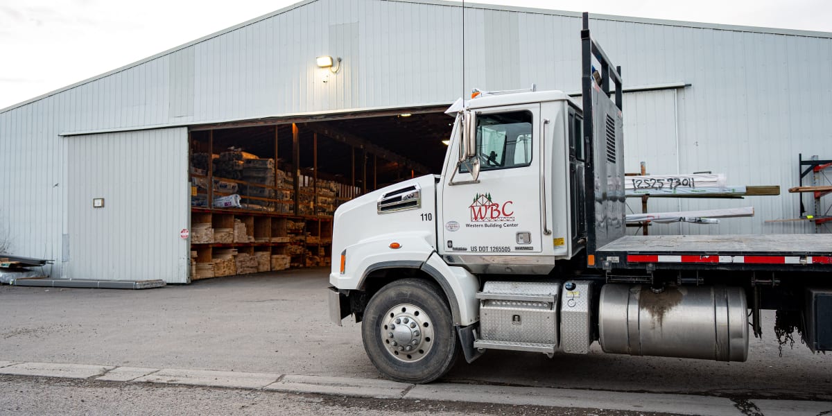 A white WBC (Western Building Center) delivery truck with flatbed trailer is parked outside a large gray metal warehouse buil