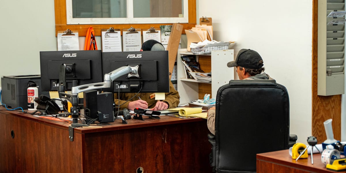 Interior office workspace showing an employee in camouflage clothing working at a wooden desk with dual ASUS monitors and var