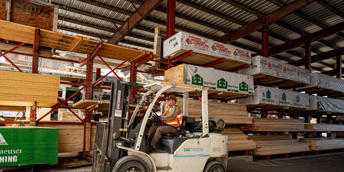 A forklift operator in an orange safety vest is working in a lumber warehouse, operating a UniCarriers Model 90 forklift near
