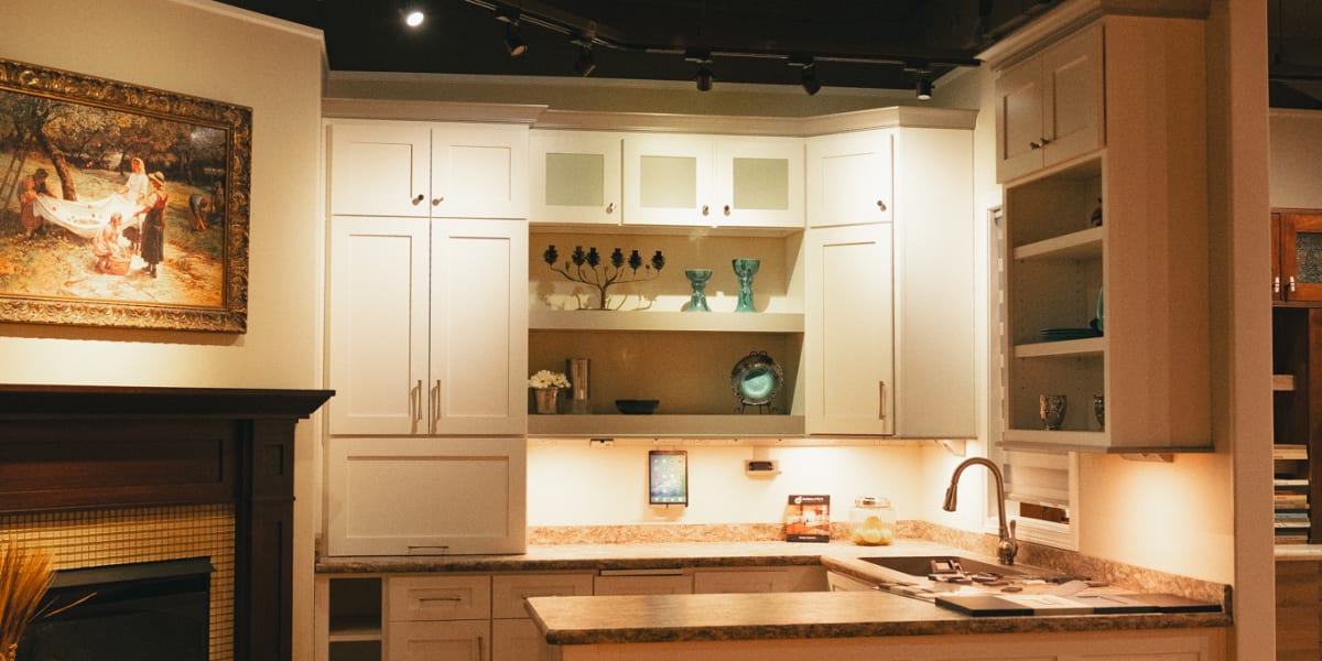 This image shows a kitchen showroom display featuring white Shaker-style cabinets with dark granite countertops and stainless