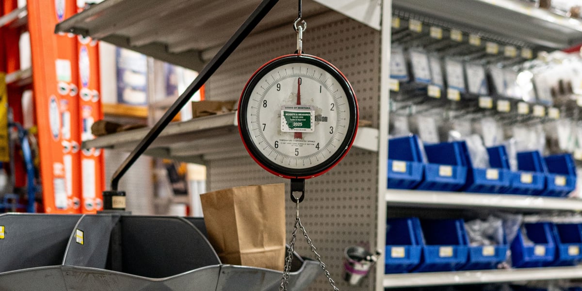 A professional hanging scale with a red rim and white dial face is suspended from a black mounting arm in a warehouse setting