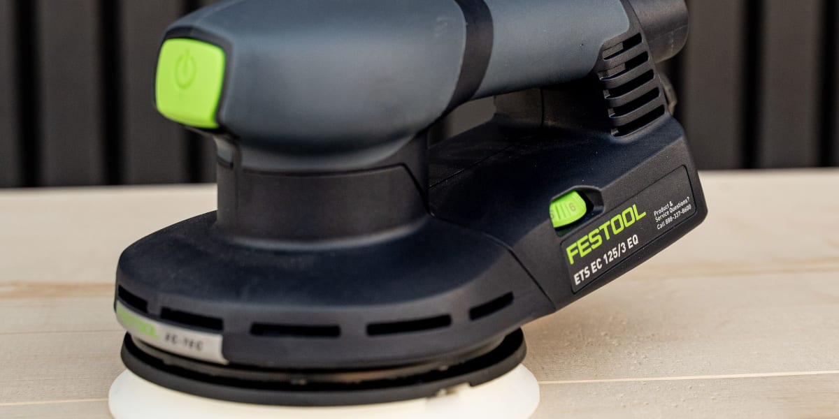 A Festool ETS EC 150/3 EQ orbital sander positioned on a light wood surface against a dark corrugated metal background