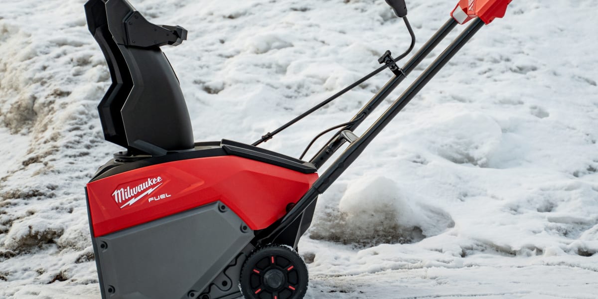 A red and black Milwaukee FUEL cordless single-stage snow blower positioned on a snowy surface with snow banks visible in the