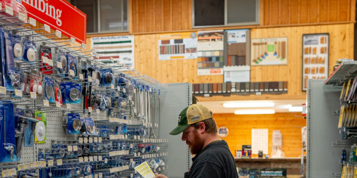 A customer in work clothes and a cap browses the plumbing aisle, reading a product package while standing among extensive she