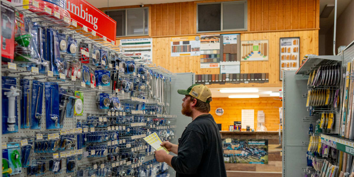 A bearded customer in a baseball cap and black jacket shops in the plumbing aisle of the store, holding what appears to be a
