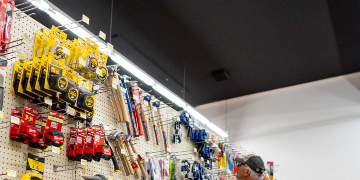 A store employee in a dark jacket and baseball cap restocks tools and hardware in the tool aisle, using a black wheeled cart