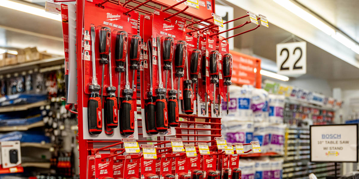 A red Milwaukee brand tool display rack in a hardware store aisle, featuring multiple tiers of screwdrivers and other hand to