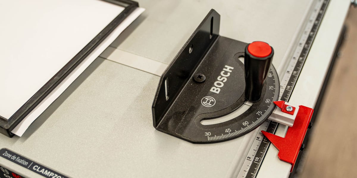 A Bosch table saw with a clear blade guard and miter gauge is shown on a white table surface