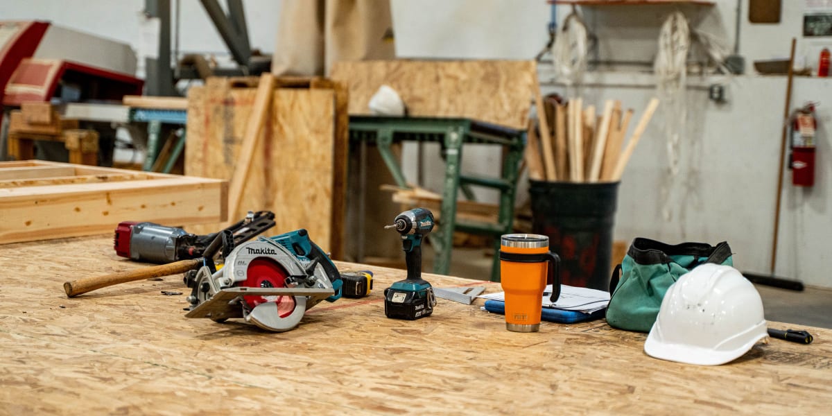 A woodworking workshop table displaying various power tools and equipment including a Makita circular saw, cordless drill wit
