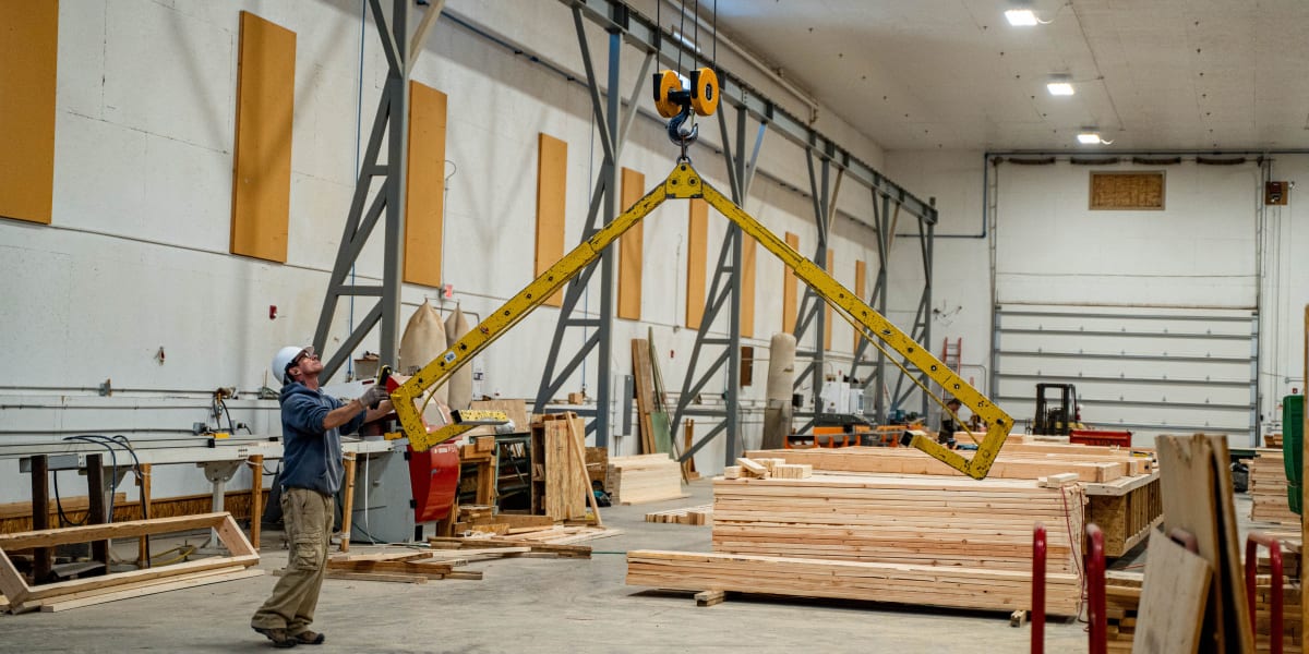 A worker operates an overhead crane system with yellow lifting spreader beam in a truss manufacturing facility