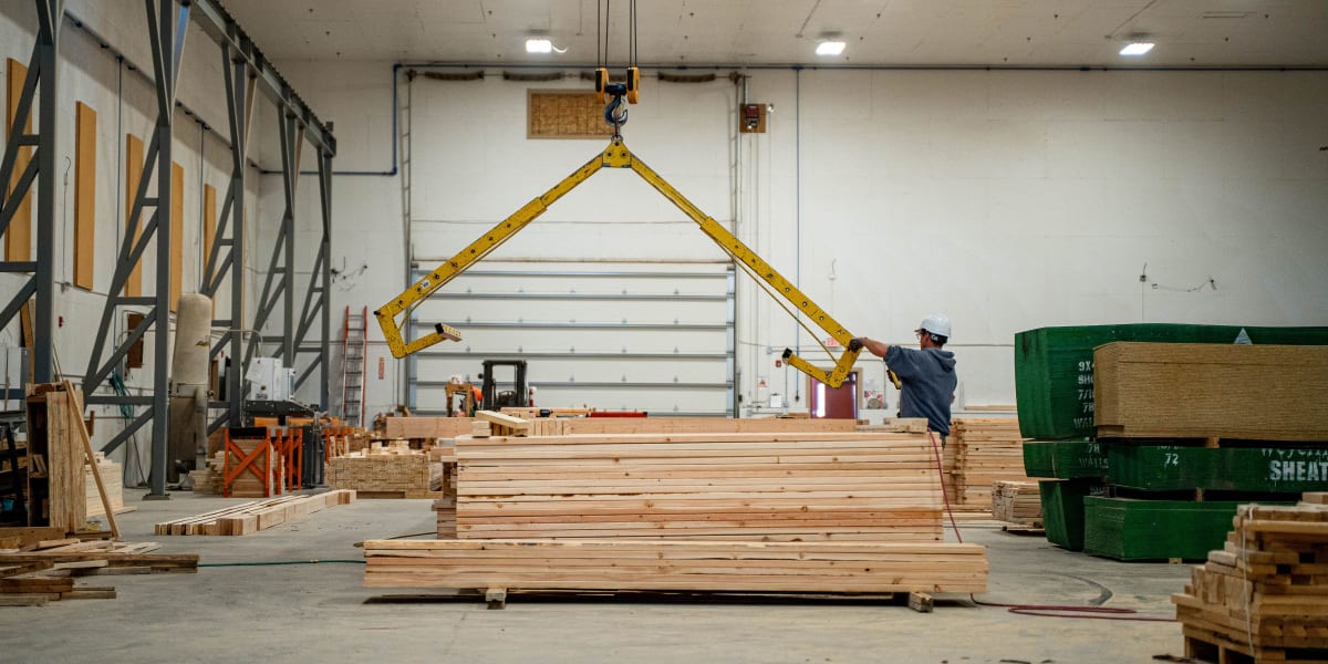 Interior view of a truss manufacturing facility showing a worker operating an overhead crane with a yellow triangular lifting