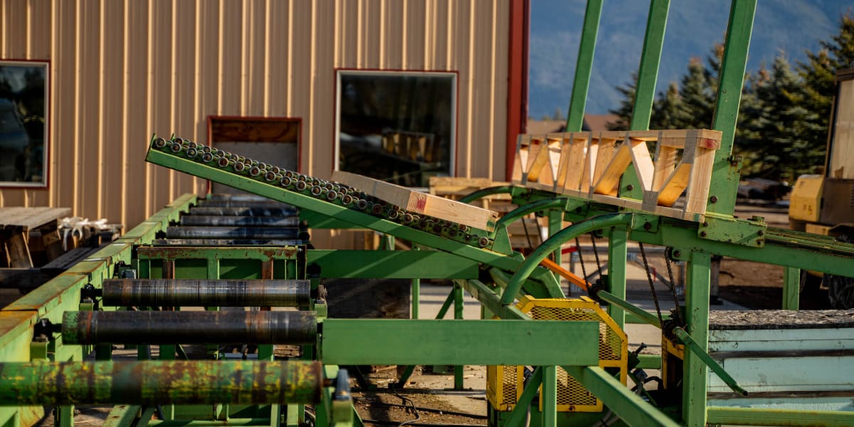 A green truss manufacturing machine positioned in front of a metal industrial building with a backdrop of snow-capped mountai