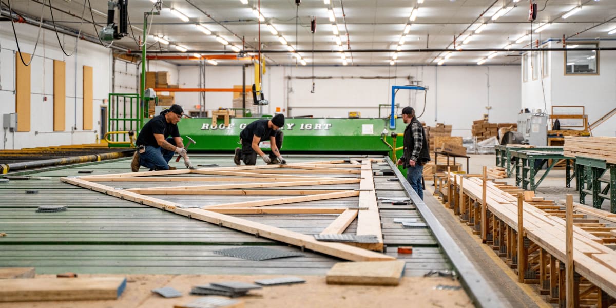 Workers in a truss manufacturing facility assembling wooden roof trusses on a large work table