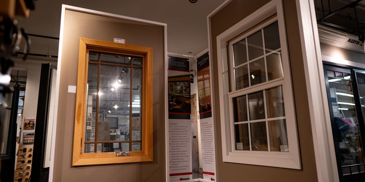 Interior showroom display featuring two window samples mounted on a gray wall - one wood-framed window on the left and one wh