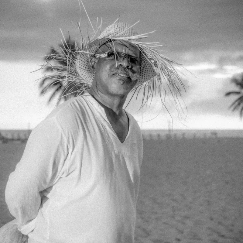 Luxury beach portrait in Santo Domingo: man with raffia accessory, gray ocean light, dark sunglasses, white texture and Caribbean wind