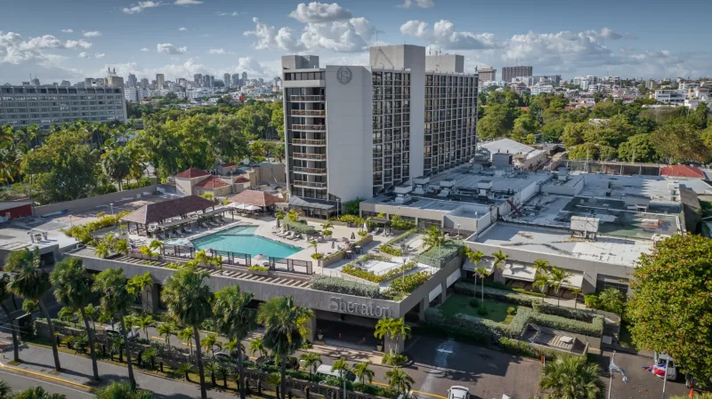 Aerial photography of residential complex in Santo Domingo: turquoise pool, white tower, palms, and urban landscape under tropical sky.