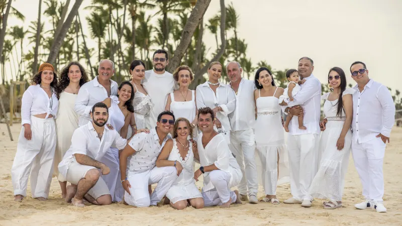 Multigenerational family photography session on Punta Cana beach, sixteen people dressed in white under palm trees with filtered natural light.