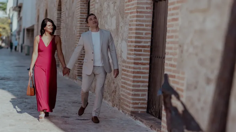 Couple walking on cobblestone streets of Santo Domingo's Colonial Zone; she wears deep red dress, he wears beige suit. Natural midday light