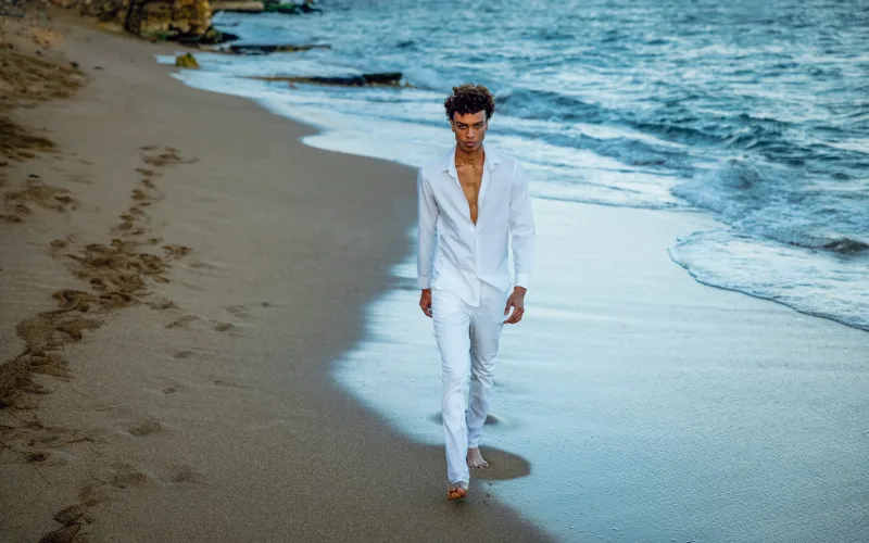 Man in fashion portrait walks barefoot through wet sand on Santo Domingo beach, wearing white linen