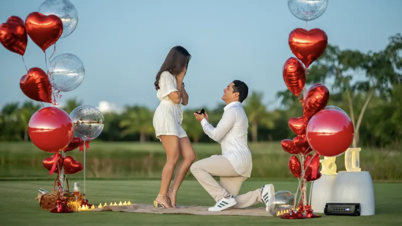 Engagement proposal in open field in Santo Domingo: man on one knee before smiling woman, surrounded by red and clear balloons, cloudless blue sky and palms in background