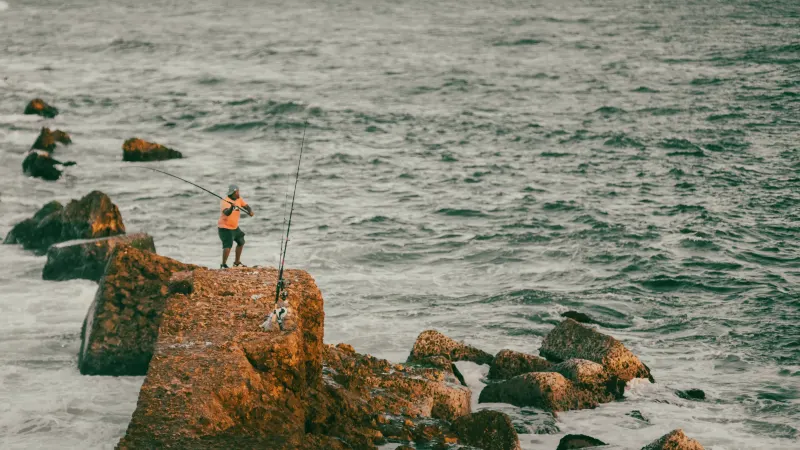 Fisherman in orange jacket standing on rust-colored rocks casting fishing rod toward sea on Dominican coast