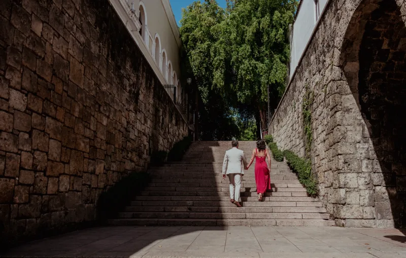 Couple holding hands walking down colonial steps in Historic Center of Santo Domingo, she wears fuchsia pink and he wears light suit