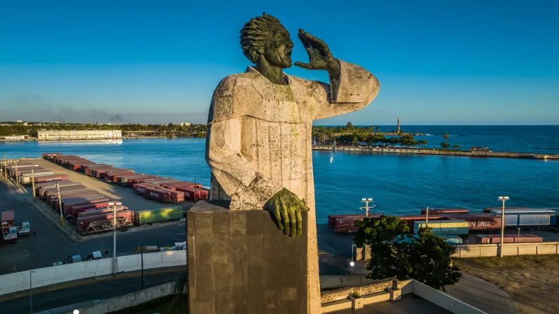 Golden stone statue with hand to cheek overlooking Santo Domingo port with containers and turquoise water, commercial photography