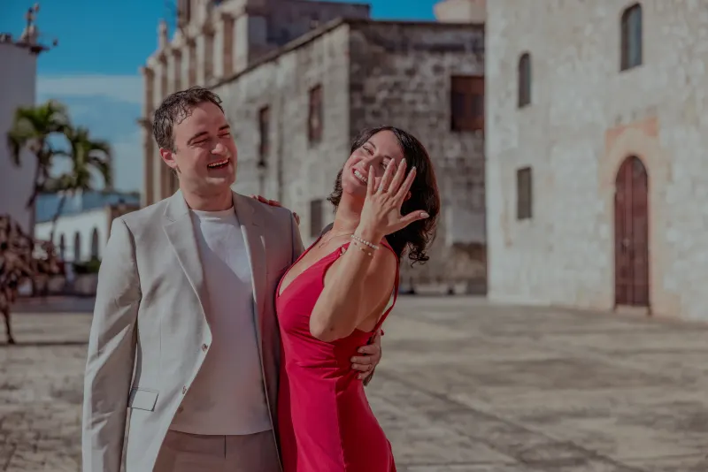 Couple laughing in Santo Domingo's colonial district plaza, woman in neon pink dress, man in beige blazer, white colonial architecture