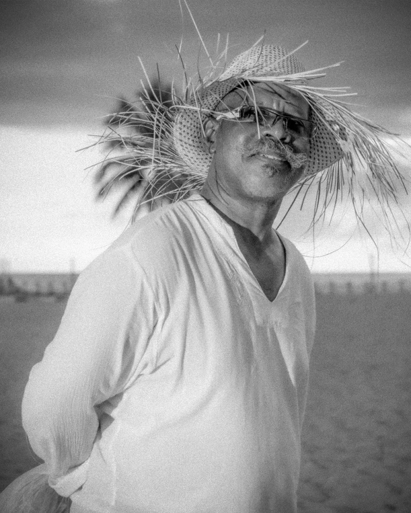 Black-and-white luxury portrait of man gazing skyward on Santo Domingo beach with royal palm and flowing white shirt
