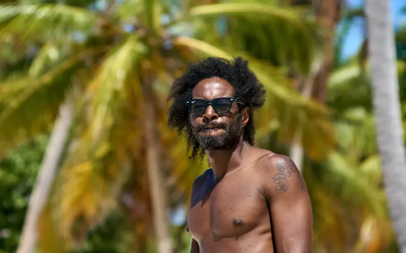 Man wearing dark sunglasses posing on tropical beach with blurred palm foliage, luxury portrait session in Punta Cana, Dominican Republic