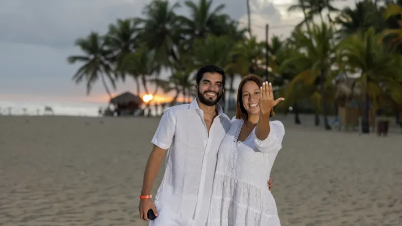 Couple in marriage proposal on Santo Domingo beach at sunset, woman showing ring with surprised smile, golden light framed by palm trees