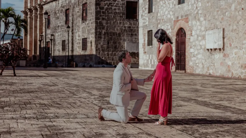Marriage proposal in Santo Domingo: man on knee at colonial plaza before woman in crimson dress, late afternoon sidelight