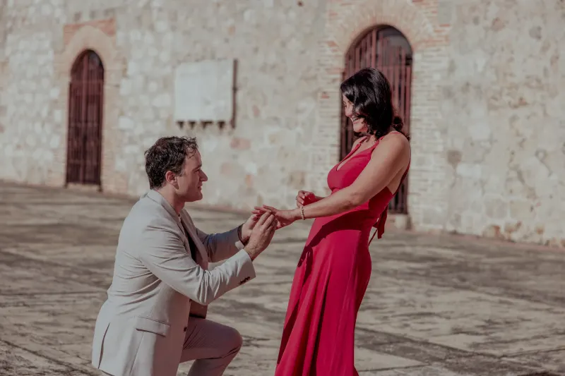 Man in beige suit kneels with ring before woman in fuchsia dress in colonial Santo Domingo plaza