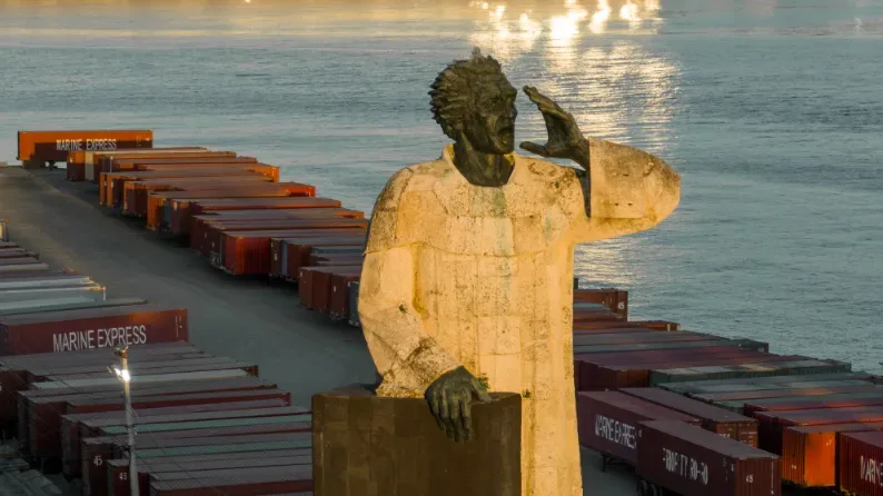 Golden statue of worker surveying cargo containers at dawn in Santo Domingo port, commercial photography with natural light.